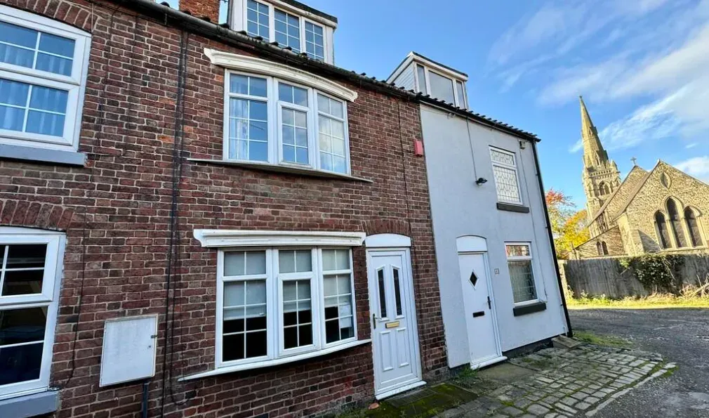 The front of 7 Ashley Terrace: a red brick terraced house with white painted bay windows and a white front door, with a stone church and spire further along the cobbled street.