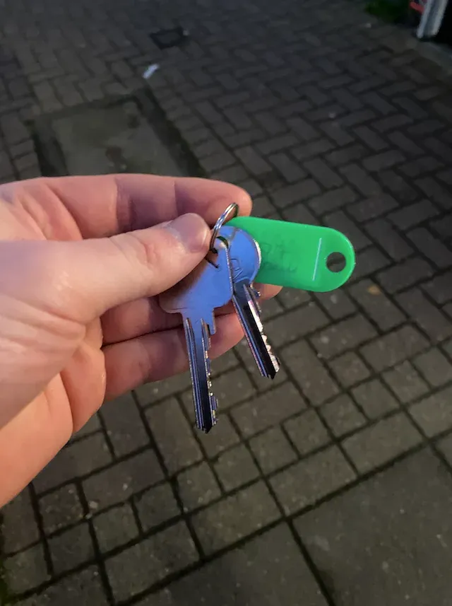 A hand holding two keys on a ring with a green plastic tag, photographed over block paving outside the property.
