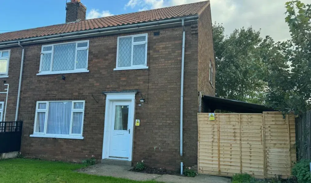 The front of 23 Beech Grove: a brown brick end of terrace house with pale blue diamond leaded windows, a white front door and a new timber fence panel along the side.