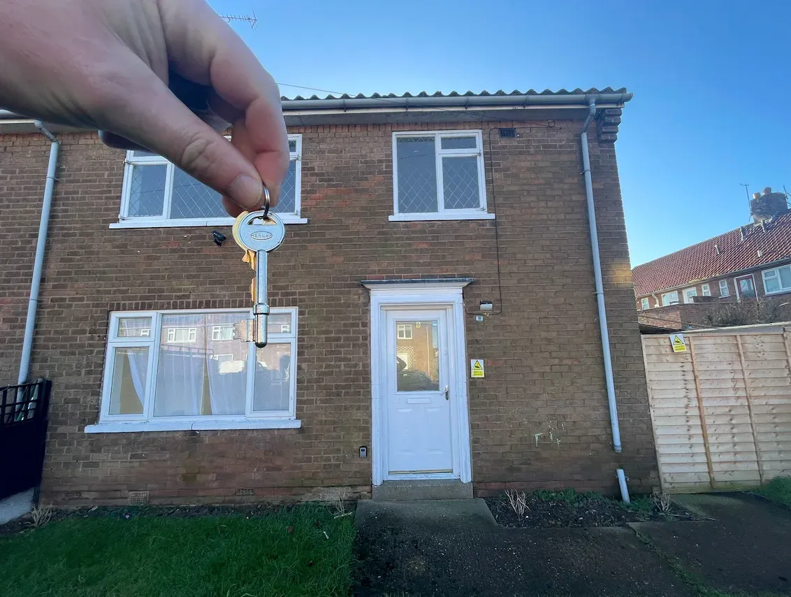 A hand holding a single key up in front of 23 Beech Grove, showing the brick end of terrace, the white front door and the front lawn under a clear blue sky.