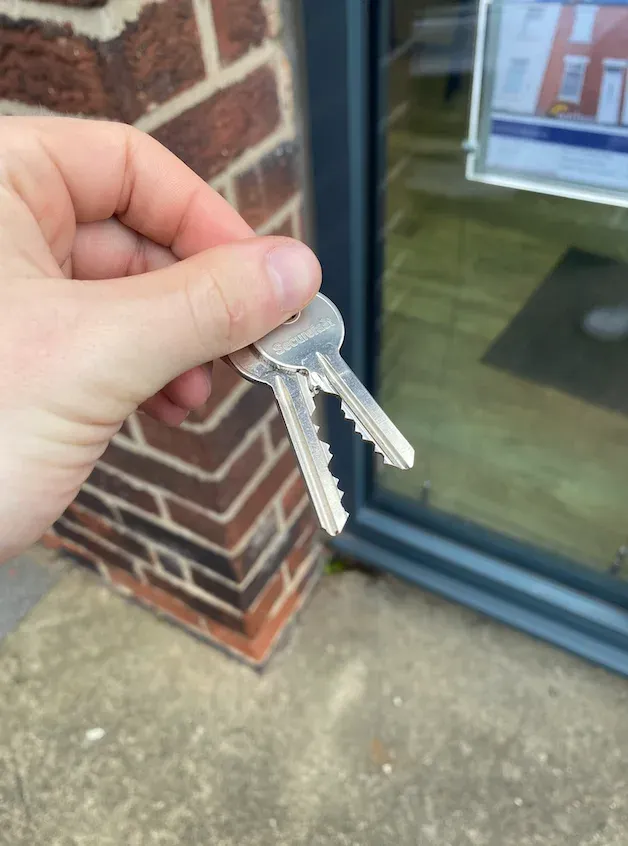 A hand holding two keys beside a brick pillar and a glazed door, with a framed property listing on display in the window behind.
