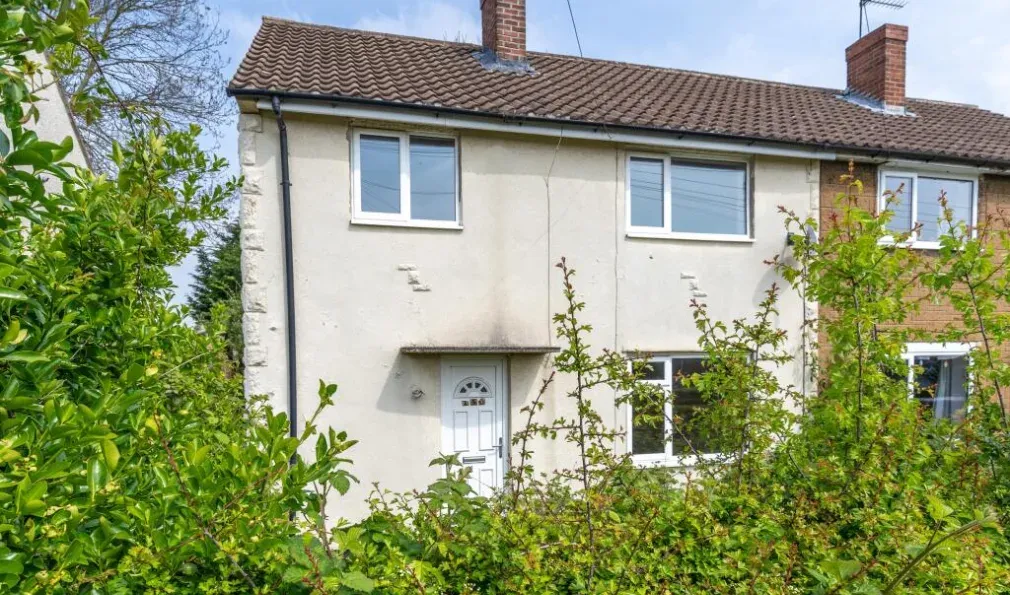 The front of 150 Long Lane: a rendered semi detached house with a brown tiled roof, white uPVC windows and a white front door, with overgrown shrubs across the front garden.