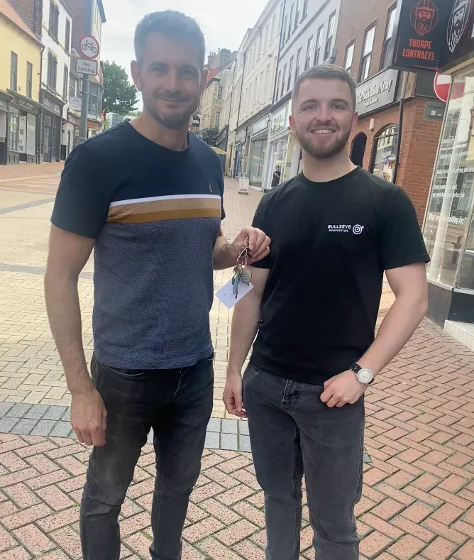 Two men standing on a pedestrianised high street, one of them in a black Bullseye Properties t-shirt, holding a set of keys with a paper tag between them.