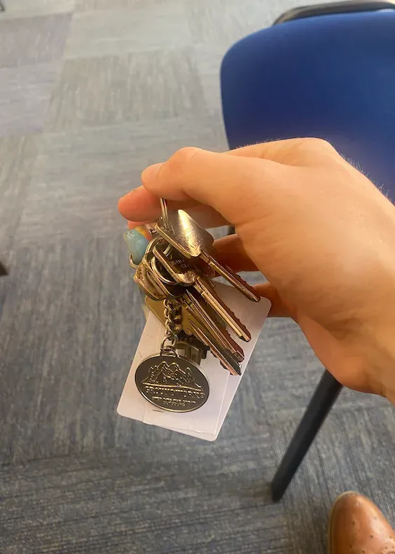A hand holding a bunch of keys with a paper tag and a round metal keyring, photographed indoors over an office carpet.