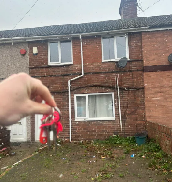 Keys on a red fob held up in sleet in front of a red brick terraced house.
