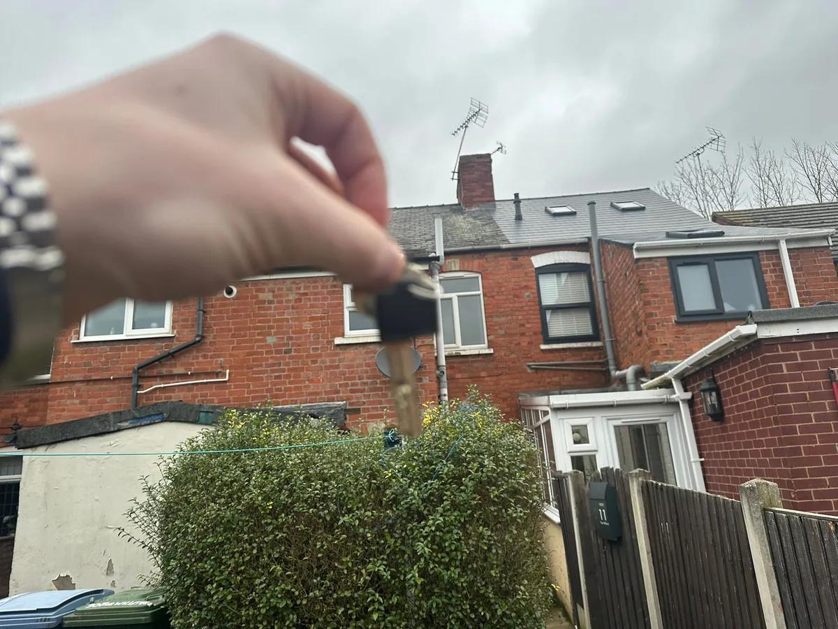 Keys held up at the rear of a red brick terraced house with a conservatory and a mature hedge.