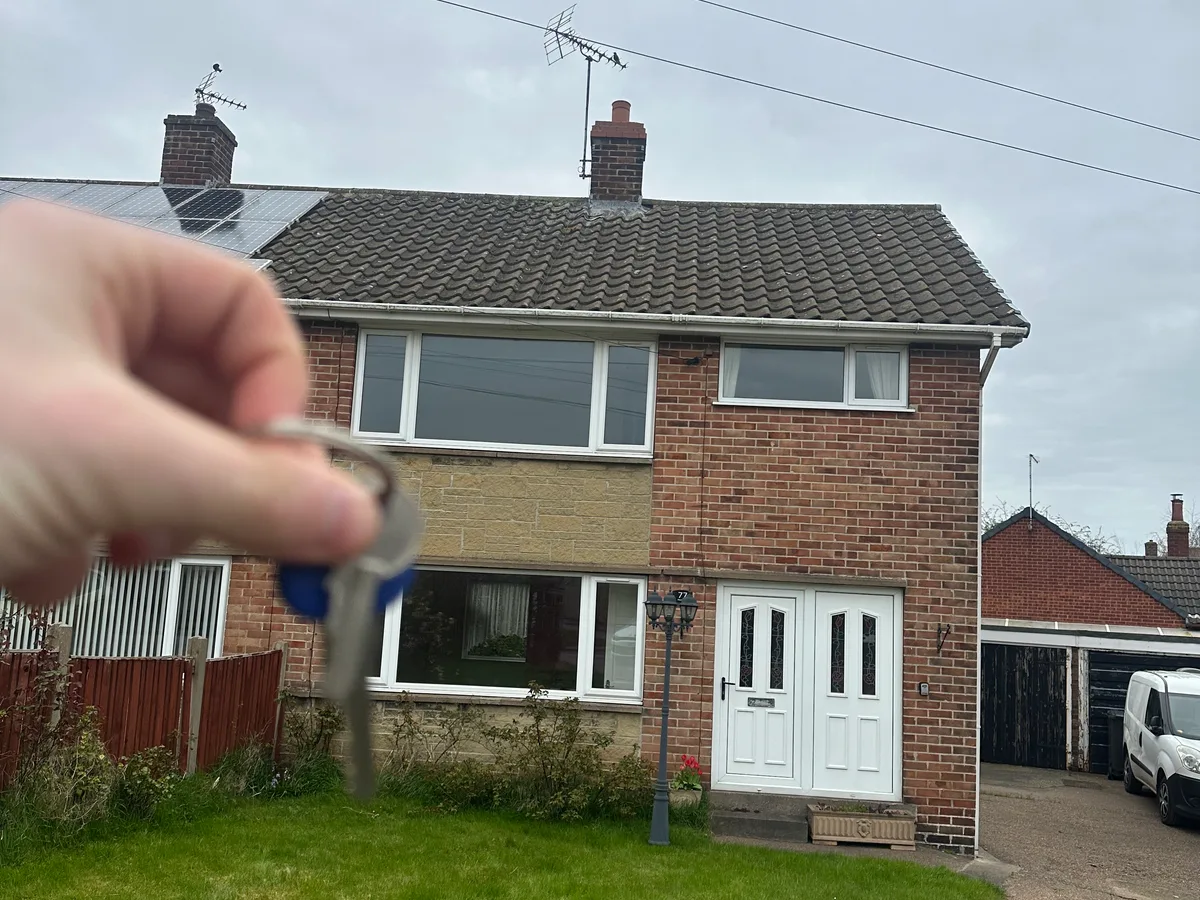 Keys held up in front of a red brick semi detached house with a white front door and solar panels on the neighbouring roof.